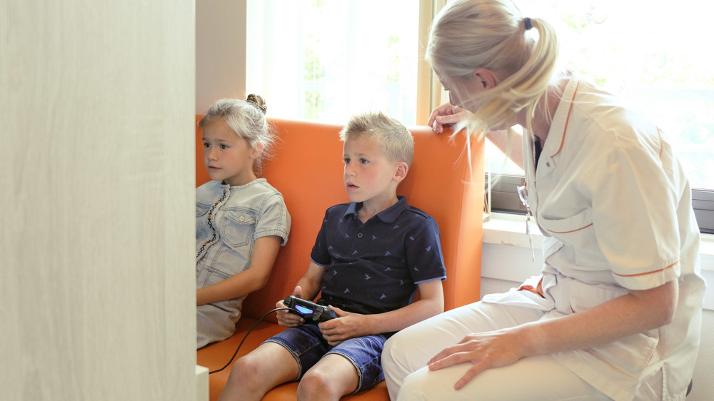 2 kinderen gamen in de speelkamer en de verpleegkundige kijkt mee.