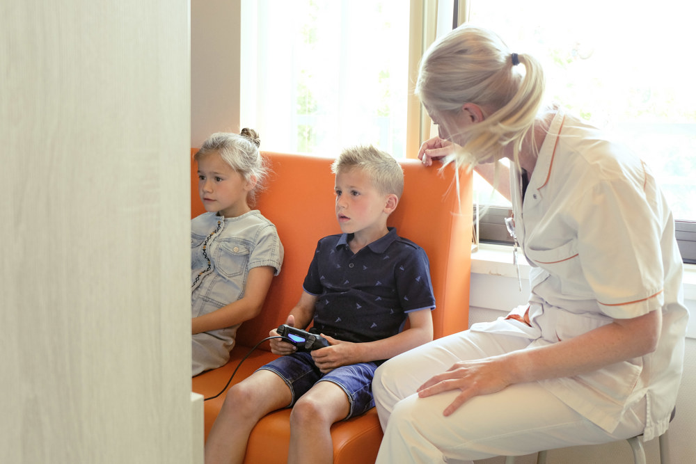 2 kinderen gamen in de speelkamer en de verpleegkundige kijkt mee.