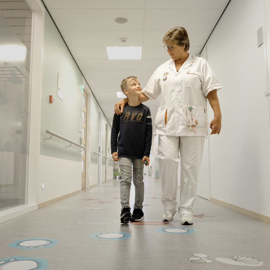 Links een kind, rechts een verpleegkundige. De verpleegkundige slaat een arm om het kind. Ze lopen in een gang.